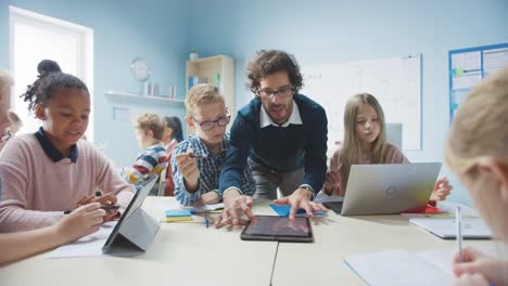 Elementary-School-Classroom:-Enthusiastic-Teacher-Holding-Tablet-Computer-Explains-Lesson-to-Brilliant-Young-Children.-Kids-Learning-Programming-Languages,-Internet-Safety-and-Information-Technology