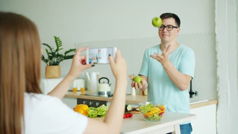 Estudiante-malabares-con-manzanas-en-la-cocina-cuando-novia-tomando-fotos-con-teléfono-inteligente