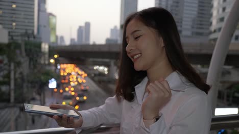 Joven-mujer-asiática-sonriente-usando-teléfono-inteligente-en-el-centro-de-la-ciudad-moderna-en-el-crepúsculo-de-Bangkok-Tailandia.-Usa-su-dedo-para-tocar-la-pantalla.-Para-usar-hologramas-Usted-diseña