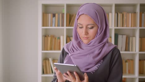 Retrato-de-primer-plano-de-joven-mujer-musulmana-en-hiyab-usando-la-tableta-y-mirando-la-cámara-de-pie-en-la-biblioteca