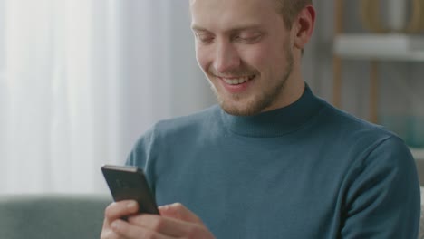 Porträt-von-hübschen-jungen-Mann-mit-Smartphone,-während-sitzen-auf-Stuhl-in-seinem-gemütlichen-Wohnzimmer.-Creative-Freelancer-entspannt-zu-Hause,-surft-im-Internet,-nutzt-Social-Media,-schaut-Videos-sendende-Nachrichten