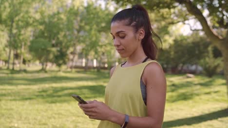 Retrato-de-una-hermosa-mujer-joven-sonriente-en-ropa-deportiva-de-pie-en-el-parque-mensajes-de-mensajes-de-texto-en-el-teléfono-inteligente