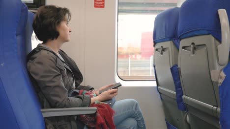 viajes-en-tren-por-ventana-mujer-caucásica-sostiene-teléfono-inteligente-Mira-por-la-ventana