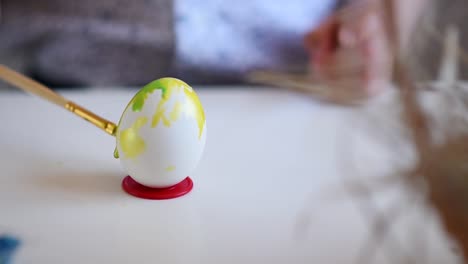 The-litlle-child-with-a-brush-paint-an-Easter-egg-in-yellow-color-on-a-stand-on-the-table.