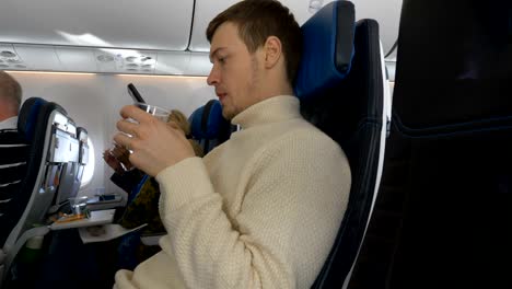 young-man-looks-at-phone-while-sitting-on-plane-drinks-water