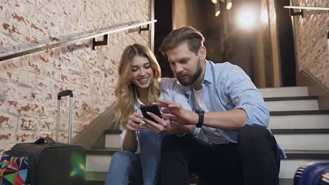 Slow-motion-of-romantic-cheerful-young-pair-which-sitting-with-their-suitcases-on-hotel's-stairs-and-using-mobile-looking-for-places-to-visit-on-holidays