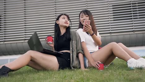 Beautiful-happy-young-asian-women-spending-time-together-near-big-modern-building-sitting-on-the-green-grass-and-talking-with