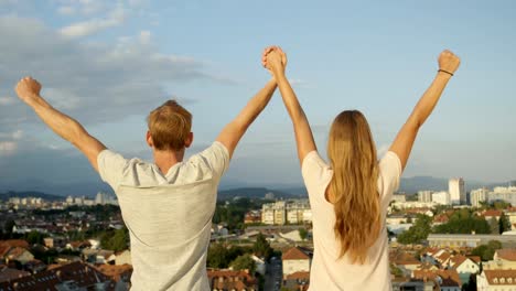 CLOSE-UP:-Girl-and-boy-standing-on-rooftop-above-the-city,-raising-hands-in-air