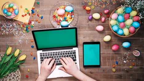 Man-using-laptop-computer-and-digital-tablet-with-green-screen-on-table-decorated-with-easter-eggs-Top-view