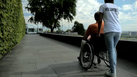 Vista-posterior-de-un-voluntariado-femenino-caminando-con-un-hombre-wheelchaired