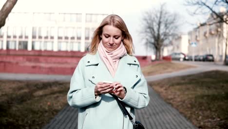 Feliz-30-años-caucásico-mujer-mecanografiar-en-el-smartphone.-Bastante-elegante-rubia-enviar-mensajes-de-texto-mientras-camina-en-la-ciudad.-Cámara-lenta