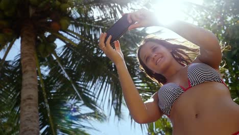 Hermosa-mujer-delgada-con-pelo-largo-morena-de-pie-junto-a-la-palmera-y-hacer-selfie-en-teléfono-móvil-en-un-cielo-azul-y-el-sol-con-fondo-de-efectos-de-la-llamarada-de-la-lente.-Chica-con-smartphone.-cámara-lenta.