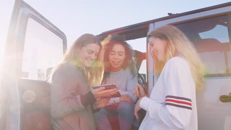 Three-young-women-on-a-road-trip-using-tablet-by-their-car