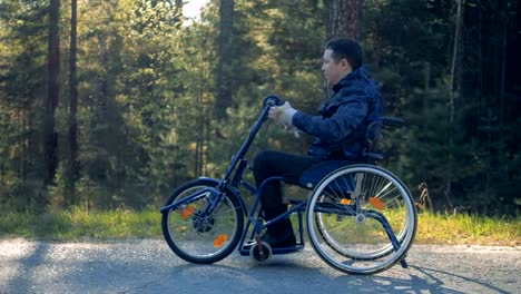 Un-hombre-con-discapacidad-va-en-una-silla-de-ruedas-en-un-camino-forestal,-vista-lateral.