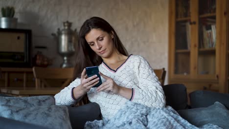 Frau-sitzt-auf-der-Couch,-eingewickelt-in-eine-Decke-und-SMS-auf-dem-Handy