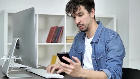 Junge-Mann-mittels-Smartphone-während-der-Arbeit-am-Desktop