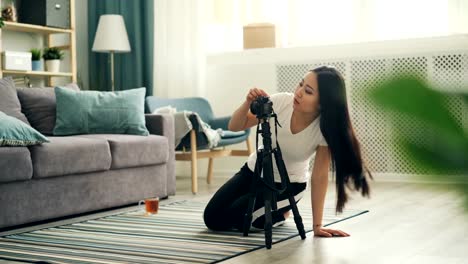 Attractive-Asian-blogger-is-installing-camera-on-tripod-and-adjusting-equipment-then-sitting-on-floor-and-recording-video-for-online-vlog.-Technology-and-lifestyle-concept.