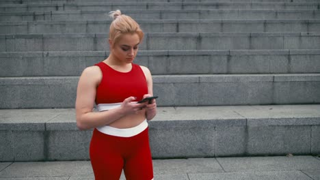 Plusgroße-blonde-gemischte-Rennracee-lächelnde-Frau-trägt-rote-Sportbekleidung-mit-Smartphone-im-Freien