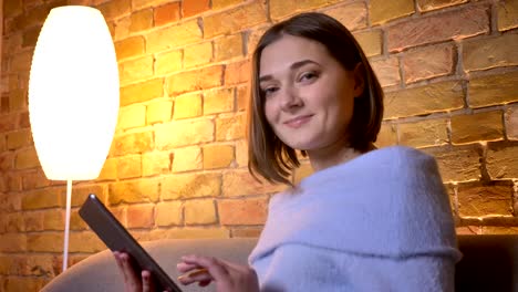 Closeup-shoot-of-young-beautiful-caucasian-female-using-the-tablet-then-looking-at-camera-and-smiling-while-sitting-on-the-couch-at-cozy-home-indoors