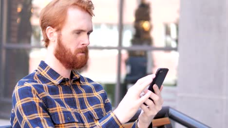 Bart-junge-Mann-mit-Smartphone-beim-Sitzen-im-Freien