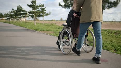 Male-is-rolling-invalid-chair-with-young-sick-woman-outdoors,-back-and-side-view