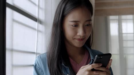 Portrait-smiling-young-asian-business-woman-holding-typing-mobile-phone-and-scrolls-through-social-media-feed-in-smartphone-standing-beside-window-at-home-office.