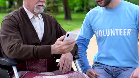 Voluntario-masculino-sonriente-explicando-el-viejo-discapacitado-cómo-utilizar-la-aplicación-del-teléfono,-cuidado