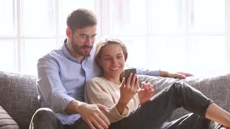 Pareja-feliz-hablar-risa-mirando-a-smartphone-sentarse-en-el-sofá