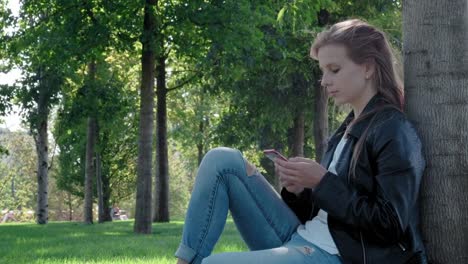 Joven-hermosa-chica-con-ropa-de-calle-en-un-parque.-Sosteniendo-un-teléfono-inteligente-en-sus-manos,-sentado-en-la-hierba-debajo-de-un-árbol.-Escribe-y-lee-mensajes-en-una-red-social.-Chatear-con-un-grupo-de-amigos