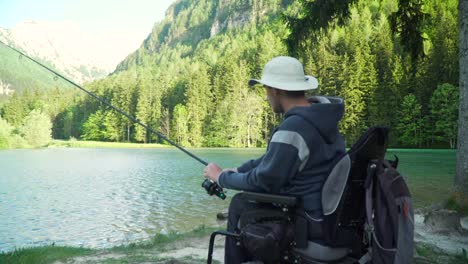 4k-Auflösung-von-behinderten-Fischer-in-einem-elektrischen-Rollstuhl-Angeln-in-schönen-See-in-der-Nähe-von-Wald-und-Berg-im-Rücken