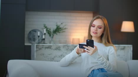 Schöne-junge-Frau-mit-fröhlichem-Ausdruck,-hält-Smartphone-und-Kreditkarte,-Banken-online-oder-macht-Einkaufen,-während-sitzt-gegen-Café-Interieur.-Zahlungs--und-Freizeitkonzept