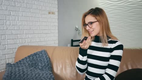 Happy-caucasian-woman-in-glasses-using-voice-recognition-on-phone-to-record-a-message-sitting-on-a-couch-at-home.-Girl-use-mobile-voice-control,-business-woman-in-modern-home