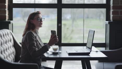 attraktive-Mädchen-hat-Spaß-im-Gespräch-auf-Video-Anruf-mit-ihrem-Freund-online-über-Laptop-zeigt-Smartphone,-während-am-Tisch-in-gemütlichen-Café-sitzen