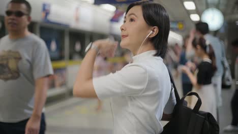 Retrato-de-mujer-asiática-con-auriculares-escuchando-música-y-usando-teléfono-inteligente-para-chatear-con-amigos-o-navegar-mientras-espera-un-tren.-Tecnología-en-la-vida-cotidiana-y-los-viajes.