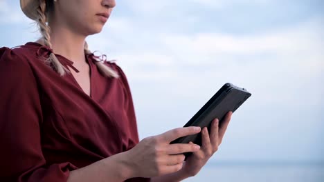 Close-up-An-attractive-young-girl-hands-in-a-summer-red-dress-sits-on-a-stone-by-the-sea-in-the-evening-and-looks-at-something-on-a-tablet.-Swipe-across-the-screen