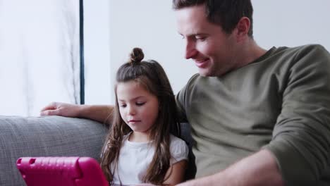 Vater-und-Tochter-sitzen-auf-Sofa-zu-Hause-spielen-zusammen-auf-digitalen-Tablet-in-rosa-Fall-zu-Hause