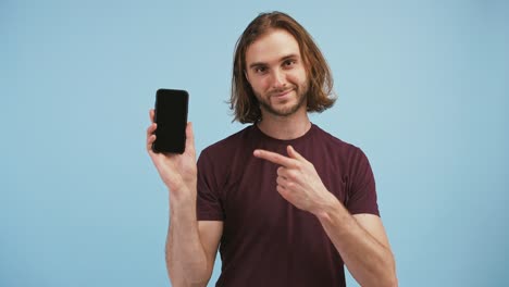 Man-raising-up-his-hand-holding-smartphone-with-black-screen,-pointing-at-it,-showing-thumb-up-and-smiling,-posing-on-blue-background