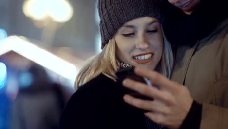 Joven-pareja-viendo-fotos-en-el-smartphone-por-la-noche-en-una-calle-de-la-ciudad.