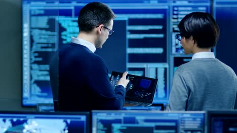 In-the-System-Control-Room-IT-Specialist-and-Project-Engineer-Have-Discussion-while-Holding-Laptop,-they're-surrounded-by-Multiple-Monitors-with-Graphics.-They-Work-in-a-Data-Center-on-Data-Mining,-AI-and-Neural-Networking.