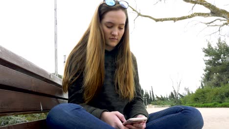 Ein-ernsthaftes-junges-Mädchen-sitzt-auf-dem-Park-Bench-mit-gekreuzten-Beinen-sitzen-und-nutzt-das-Telefon.