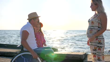 child-And-Dad-hold-to-mothers-belly,-man-in-wheelchair-with-little-Girl-and-pregnant-woman-outdoors