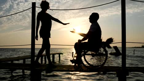 silhouette-of-loving-couple-with-disabled-person-on-seafront-near-shiny-water-in-bright-beams