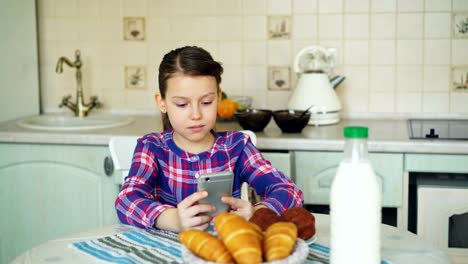 Little-beautiful-girl-playing-smartphone-at-morning-while-sitting-at-table-in-kitchen.-Childhood,-people,-and-technology-concept