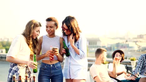 Joyful-female-friends-are-using-smartphone-and-chatting-holding-bottles-during-party-on-roof.-Social-media,-young-people-and-modern-technology-concept.