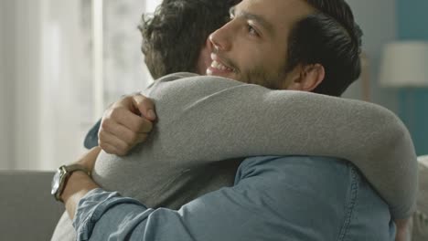 Pareja-Gay-masculino-atractivo-lindo-sentarse-juntos-en-un-sofá-en-casa.-Novios-son-Hugging-y-abrazándose-unos-a-otros.-Ellos-son-feliz-y-sonriente.-Son-vestidos-casualmente-y-sala-tiene-un-Interior-moderno.