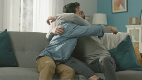 Cute-Attractive-Male-Gay-Couple-Sit-Together-on-a-Sofa-at-Home.-Boyfriends-are-Hugging-and-Embracing-Each-Other.-They-are-Happy-and-Smiling.-They-are-Casually-Dressed-and-Room-Has-Modern-Interior.