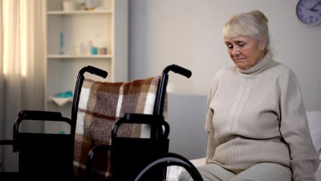 Upset-elderly-woman-sitting-on-sofa-near-wheelchair-in-rehabilitation-center