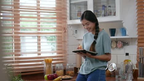 Mujer-asiática-joven-usando-la-tableta-y-comiendo-pan.-tecnología,-red-social,-concepto-de-comunicación-en-la-cocina-en-casa.