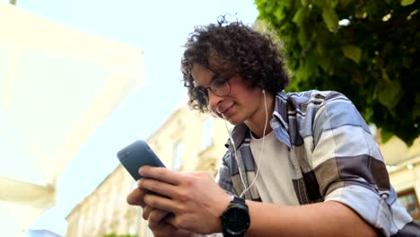 Lächelnd-schmungend-Mann-in-Kopfhörern-per-Smartphone,-Musik-hören-und-auf-Smartphone-surfen.-Outdoor.-Close-up.-Blogger-scrollt-durch-soziale-Medien-auf-Gerät,-lesen-Nachrichten-auf-App