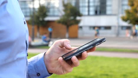 Cerrar-mano-de-joven-empresario-sosteniendo-y-tocando-el-teléfono-inteligente.-Hombre-irreconocible-parado-en-la-calle-urbana-y-el-teléfono-de-navegación.-Un-tipo-usando-artilugio-para-trabajar.-Vista-lateral-de-cámara-lenta